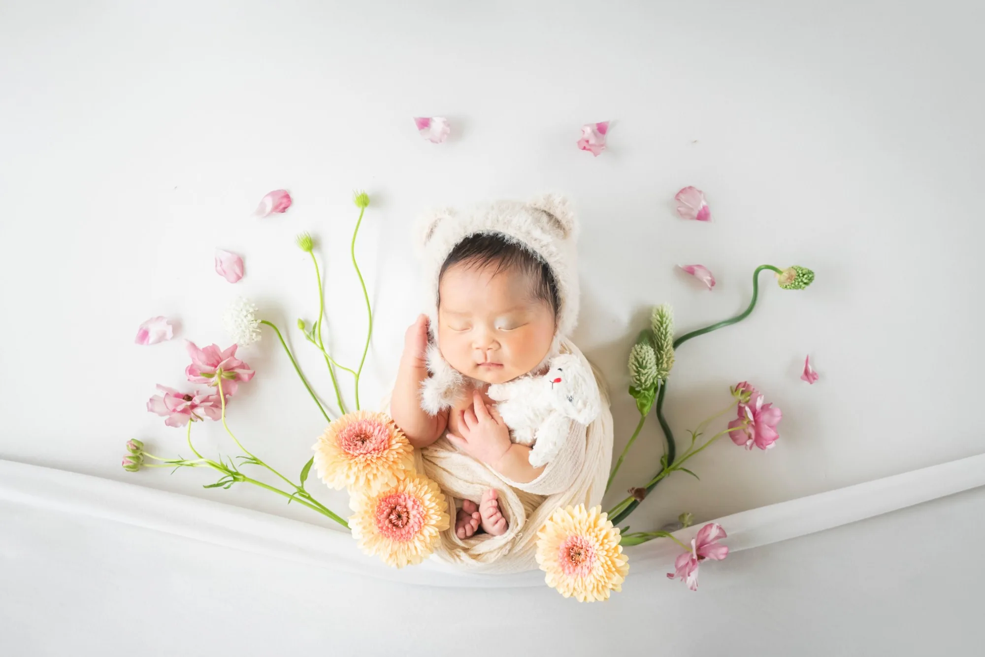 くまの帽子をかぶり、花に囲まれて眠る新生児
